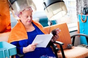 Older female customer reading a magazine in a hair salon, under a dryer with orange towel.